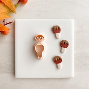 May include: A white square tile displays a cookie cutter and three miniature apple-shaped biscuits. The cookie cutter is light orange, and the biscuits are red with brown tops and white sticks. Autumn leaves and berries are in the upper left corner.