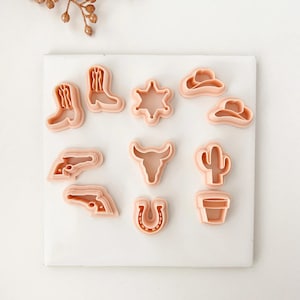 May include: A selection of light pink cookie cutters in Western-themed shapes. The cutters include cowboy boots, a sheriff's star, cowboy hats, a bull's head, a cactus, a horseshoe, and pistols. They are arranged on a white surface.