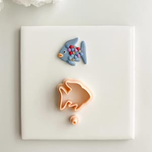 Peut inclure: Une figurine de poisson bleue faite à la main avec des accents colorés, à côté d'un emporte-pièce en forme de poisson. Le poisson a un corps rond, une petite bouche orange et une queue. L'emporte-pièce est de couleur pêche.