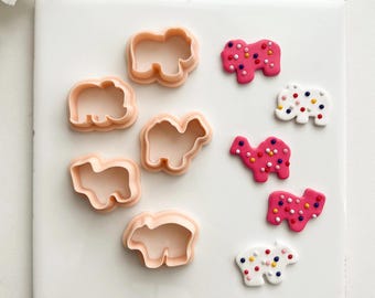 May include: A collection of light peach-colored animal-shaped cookie cutters, including elephants, camels, and hippos, alongside decorated cookies. The cookies are pink and white, adorned with colorful sprinkles. The items are arranged on a white surface.
