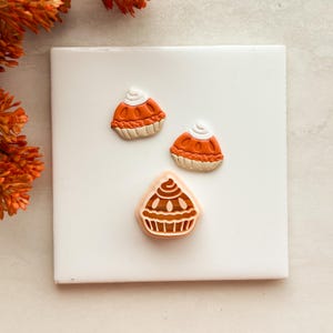 May include: Two small orange and white pie-shaped charms with a dollop of cream on top, alongside a light peach-coloured plastic cookie cutter featuring a detailed cupcake or pie design with a scalloped base and a swirled top.
