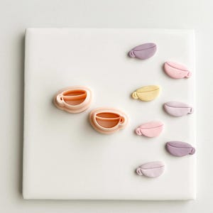 Puede incluir: Una colección de decoraciones en forma de taza de café en colores pastel. Las tazas son en tonos de rosa, amarillo y morado, dispuestas sobre una superficie cuadrada blanca. El diseño es simple y minimalista, con un enfoque en el color y la forma.