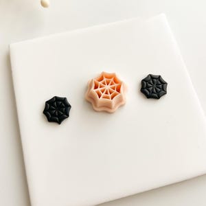 May include: Three spiderweb-shaped objects. Two are black, and one is peach-colored. The objects are arranged on a white surface. The spiderweb designs are detailed and symmetrical, suggesting a Halloween or spooky theme.