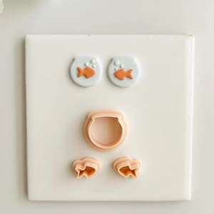 Puede incluir: Un conjunto de cortadores de galletas de color melocotón claro con forma de pecera, pez y burbujas. Dos galletas de pecera con peces naranjas en la parte superior. Un cortador de pecera en el medio y dos cortadores de peces en la parte inferior.