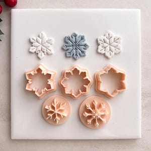 May include: A collection of snowflake-shaped cookie cutters and stamps in various designs. The cutters are a light peach color, while the stamped snowflakes are white and blue with gold accents. The items are arranged on a white surface.