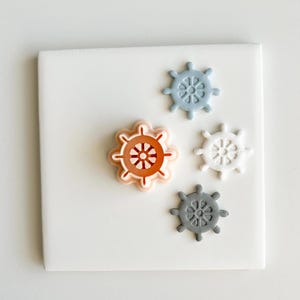 Pode incluir: Quatro objetos em forma de leme em tons de laranja, cinza, branco e azul estão dispostos em um quadrado branco. A maior roda é laranja com um centro laranja mais claro. Os outros três são menores e em diferentes tons de cinza, branco e azul.