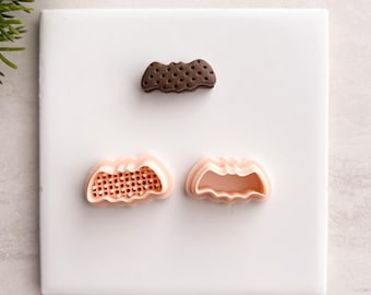 May include: A top-down view of three cookie cutters on a white surface. One is a dark brown bat shape with small holes. The other two are light pink pumpkin shapes, one with a grid pattern inside.