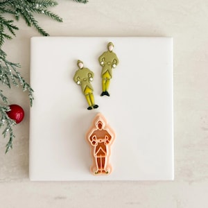 May include: Three decorative soldier-shaped items. Two are painted with olive green coats, yellow pants, and black shoes. The third is a cookie cutter in the shape of a soldier. The items are on a white square surface with a sprig of greenery and a red ornament.