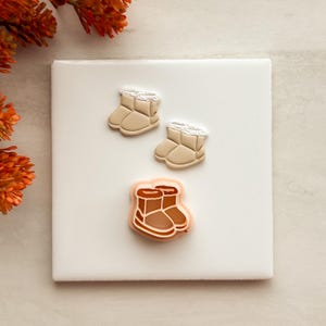 Puede incluir: Dos pequeños dijes con forma de bota en color beige con borde blanco texturizado y un cortador de galletas más grande de color melocotón con forma de par de botas y detalles interiores marrones, sobre una superficie blanca cuadrada.