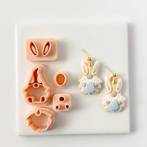 May include: A collection of pink polymer clay cutters in various shapes, including bunny ears, a gnome, and a beard. Two finished Easter bunny earrings with yellow bodies, white fur, and blue polka dot eggs are also present.