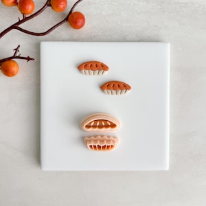 May include: Miniature polymer clay pie shapes in orange and beige, arranged on a white surface. The pieces include two complete pies and two pie crusts. A branch with orange berries is in the background.
