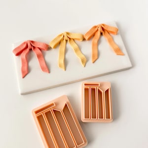 Puede incluir: Tres adornos en forma de lazo en coral, amarillo y naranja sobre una superficie rectangular blanca. También se ven dos cortadores de galletas melocotón, con un diseño rectangular y líneas verticales.