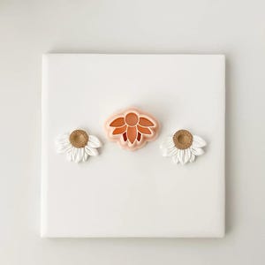 Puede incluir: Un lienzo blanco muestra tres diseños florales. Dos son blancos con centros beige, y uno es un cortador de galletas con forma de flor melocotón y naranja. Los diseños están dispuestos horizontalmente, sugiriendo un tema de artesanía o repostería.