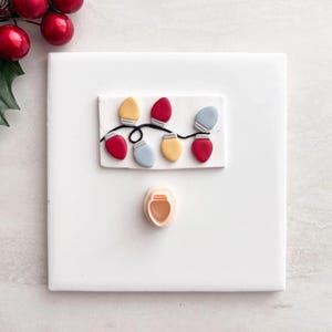 May include: A white square with a decorative string of Christmas lights in red, yellow, and blue. A small, peach-colored cookie cutter is below the lights. Red berries and green leaves are in the upper left corner.