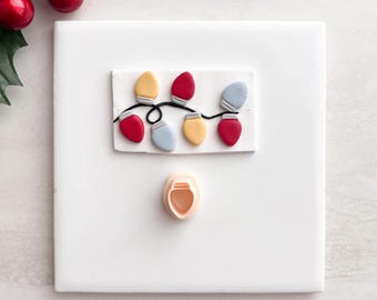 May include: A white square with a decorative string of Christmas lights in red, yellow, and blue. A small, peach-colored cookie cutter is below the lights. Red berries and green leaves are in the upper left corner.