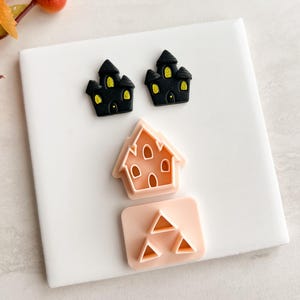 May include: Halloween-themed cookie cutters. Two black castle cutters with yellow windows and doors, a peach-colored house cutter, and a peach-colored cutter with three triangles. The cutters are on a white surface.