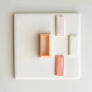 Puede incluir: Una baldosa cuadrada blanca con cuatro objetos rectangulares en tonos rosa y blanco. Un objeto es un marco hueco, y los otros son rectángulos sólidos. Los objetos están dispuestos sobre la baldosa.