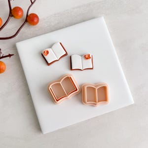 May include: A collection of book-shaped cookie cutters and decorated cookies. The cookies have white pages, brown borders, and orange flower accents. Two cutters are open books, and two are the cutter outlines.