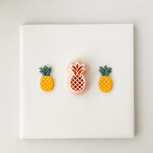 May include: Three pineapple-shaped decorations on a white background. Two are yellow with green tops, and the center one is pink with a red interior. The pineapples have a textured surface.
