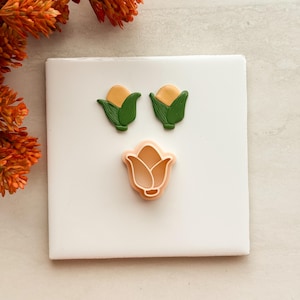 Peut inclure: Deux petites boucles d'oreilles en forme de maïs avec des grains jaunes et des feuilles vertes, accompagnées d'un emporte-pièce en plastique pêche en forme de maïs. Les objets sont disposés sur une surface carrée blanche.