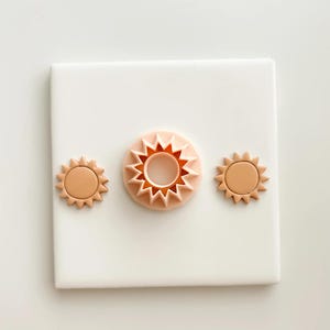 May include: Three sun-shaped cookie cutters in varying sizes. The cutters are a light peach color and feature a circular center with pointed rays extending outwards. The cutters are arranged on a white square surface, creating a clean and minimalist aesthetic.