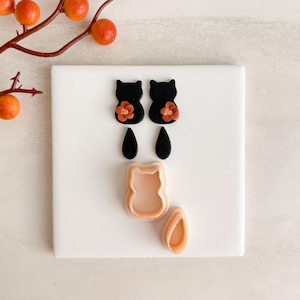 May include: A collection of black cat-shaped earrings with orange flower accents, black teardrop-shaped earrings, and cat-shaped and teardrop-shaped clay cutters. The items are arranged on a white surface, with orange berries in the background.