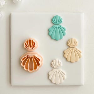 May include: A collection of seashell-shaped cookie cutters in various sizes and colors, including peach, teal, and cream. The cutters are arranged on a white surface, showcasing their intricate designs. Ideal for creating beach-themed treats.