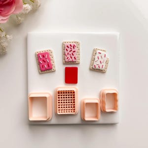 May include: A collection of polymer clay food miniatures, including three miniature Pop-Tarts with pink, red, and white frosting and sprinkles. Also included are several square-shaped cookie cutters in shades of peach and red.