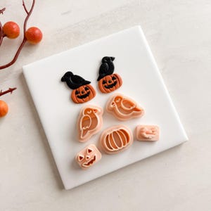 May include: A selection of Halloween-themed cookie cutters. The set features orange pumpkins with carved faces, black crows, and bird-shaped cutters. The cutters are displayed on a white surface, ideal for making festive treats.