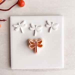 Puede incluir: Una baldosa cuadrada blanca con tres decoraciones blancas en forma de libélula y un cortador de galletas en forma de libélula de color melocotón. La baldosa está sobre una superficie clara, con una rama de bayas rojas en la esquina superior izquierda.