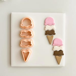 Puede incluir: Un juego de cuatro cortadores de galletas de plástico color melocotón con forma de bolas y conos de helado. También se muestran dos conos de helado terminados con bolas rosas, blancas y marrones. Los artículos están dispuestos sobre una superficie blanca.