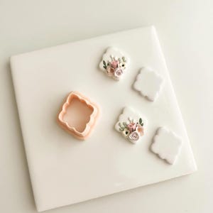 May include: A light pink cookie cutter and three white, floral-decorated biscuits. The biscuits feature pink, cream, and green floral designs. Two additional blank biscuits are also present. The items are arranged on a white surface.