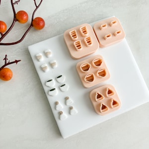 May include: A selection of miniature food-themed cookie cutters and decorative elements. The cutters are arranged on a white surface, with shapes including coffee cups, to-go cups, and winter hats. A branch with orange berries is in the background.