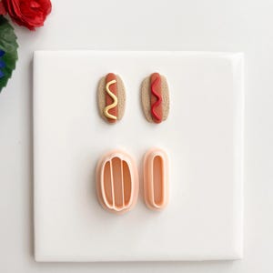 Puede incluir: Un juego de cortadores de arcilla polimérica con temática de perritos calientes y dos miniaturas de perritos calientes terminados. Una miniatura tiene mostaza amarilla, la otra tiene ketchup rojo. Los cortadores son de plástico color melocotón.