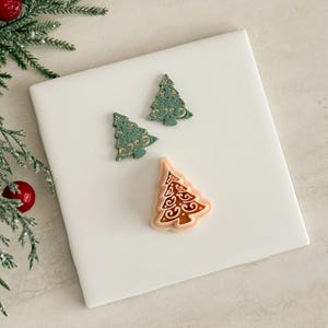 Puede incluir: Tres adornos de árbol de Navidad. Dos son verdes con detalles dorados, y uno es melocotón con detalles marrones. Los adornos están sobre una superficie blanca, con una ramita de hoja perenne y una bola roja en el fondo.