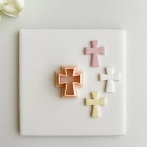 May include: A collection of cross-shaped cookie cutters in various colors, including pink, white, and yellow, arranged on a white surface. One cutter is a light peach color. The crosses are likely for baking or crafting.
