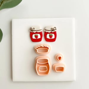 May include: A collection of pink plastic cookie cutters shaped like a jar of strawberry jam. Two assembled jars are red with white lids and a strawberry graphic. The set includes jar, lid, and strawberry shapes.
