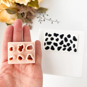 May include: A hand holds a light pink cookie cutter with six different shapes. Beside it is a white square with a black and white cow print design. The background includes yellow flowers and greenery.