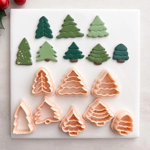 以下が含まれることがあります： さまざまな緑と桃色のクリスマスツリー型のクッキーカッターとデコレーションクッキーの詰め合わせ。クッキーとカッターは白い表面に配置され、右上隅には赤いベリーの小枝があります。