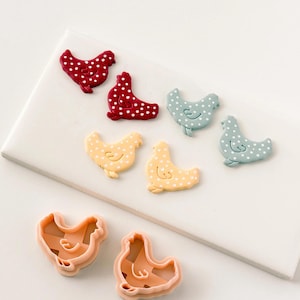 May include: A collection of chicken-shaped items in various colors. There are red, light blue, and yellow chickens with white polka dots. Two peach-colored chicken-shaped cookie cutters are also present. The items are arranged on a white surface.