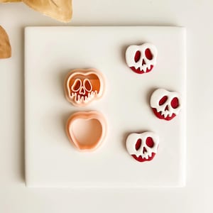 Peut inclure: Ensemble de découpoirs à biscuits et éléments décoratifs sur le thème d'Halloween. Les découpoirs représentent une tête de mort et une pomme. Les éléments décoratifs sont blancs avec des accents rouges, ressemblant à un crâne. Sur une surface blanche.