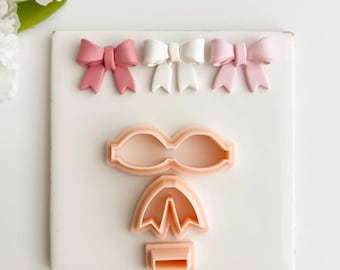 May include: Three decorative bows in pink and white are arranged above three cookie cutters. The cookie cutters are in the shape of a bow, an umbrella, and a rectangular base. The items are on a white surface.