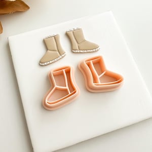 May include: Two pairs of cookie cutters shaped like winter boots. One pair is a light peach color, and the other pair is tan with a white trim. The cutters are on a white surface.