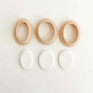 May include: Three oval-shaped cookie cutters in a light peach color are arranged above three white, textured oval shapes. The cutters are made of plastic and have a smooth finish. The white shapes appear to be made of a textured material.