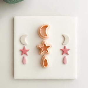 Puede incluir: Un conjunto de cortadores de galletas rosas y blancos con formas celestiales, incluyendo lunas, estrellas y lágrimas, dispuestos sobre una superficie blanca. Los cortadores son de plástico y probablemente se utilizan para hornear o manualidades.