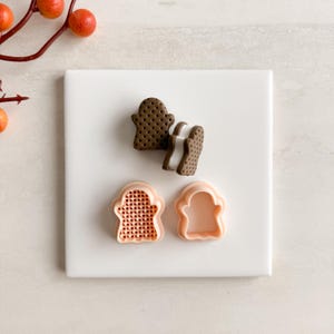 May include: A top-down view of a white square tile with a ghost-shaped cookie and cookie cutters. The cookie is brown with a dotted pattern, and the cutters are peach-colored. A sprig of orange berries is in the upper left corner.