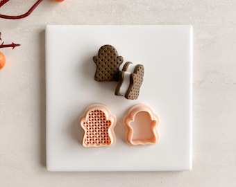 May include: A top-down view of a white square tile with a ghost-shaped cookie and cookie cutters. The cookie is brown with a dotted pattern, and the cutters are peach-colored. A sprig of orange berries is in the upper left corner.