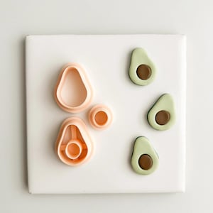 Puede incluir: Un conjunto de cortadores de galletas con forma de aguacate y alimentos con forma de aguacate. Los cortadores son de plástico color melocotón, y las mitades de aguacate son verdes con centros marrones. Los artículos están dispuestos sobre una superficie blanca.