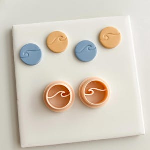 Puede incluir: Un conjunto de cortadores de galletas y elementos decorativos redondos con forma de ola. Los cortadores son de color melocotón, mientras que los elementos decorativos son azules y beige. Los cortadores y las piezas tienen un diseño de ola.