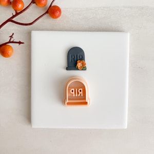 Könnte beinhalten: Ein dunkelgrauer Grabstein mit den Buchstaben "RIP" und einer kleinen orangefarbenen Blume. Darunter befindet sich ein pfirsichfarbener Ausstecher in Form eines Grabsteins mit den Buchstaben "RIP" in umgekehrter Reihenfolge. Halloween-Thema.